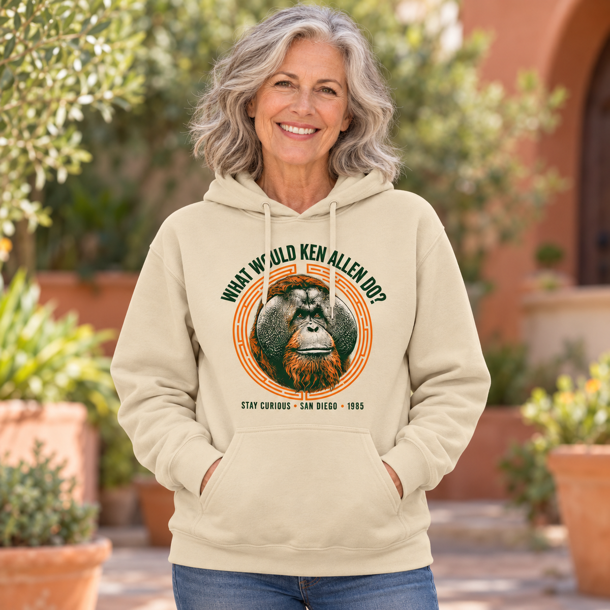 Sand What Would Ken Allen Do? pullover hoodie with green lettering and orange orangutan artwork in a leafy terracotta courtyard