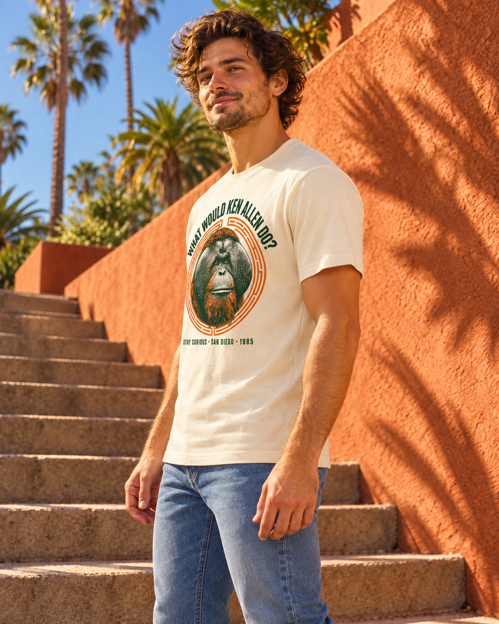 Three-quarter side view of the Natural What Would Ken Allen Do? tee with orange orangutan artwork beside a terracotta wall